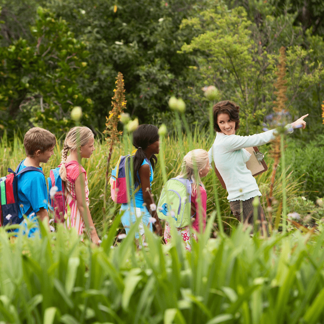 Children with backpacks walk in a line on a green nature trail. A female guide leads, pointing out something in the distance. Bright, sunny scene.