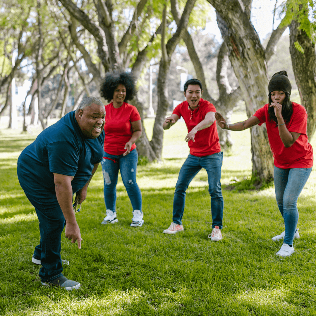 Four adults in a park doing a team-building activity. One bends down, laughing, while three in red shirts cheer. Tall trees and green grass create a lively scene.