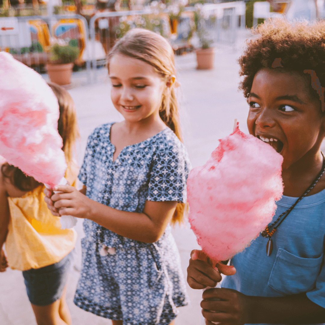 Three children at a fair with pink cotton candy. A curly-haired boy eagerly bites his candy, while a girl in a blue dress and another child smile.