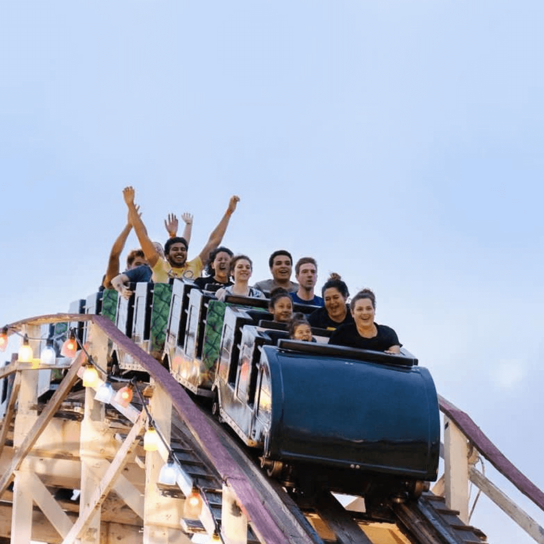 People on a roller coaster at the peak, some with hands raised, others gripping safety bars. Clear sky emphasizes the thrill and height.